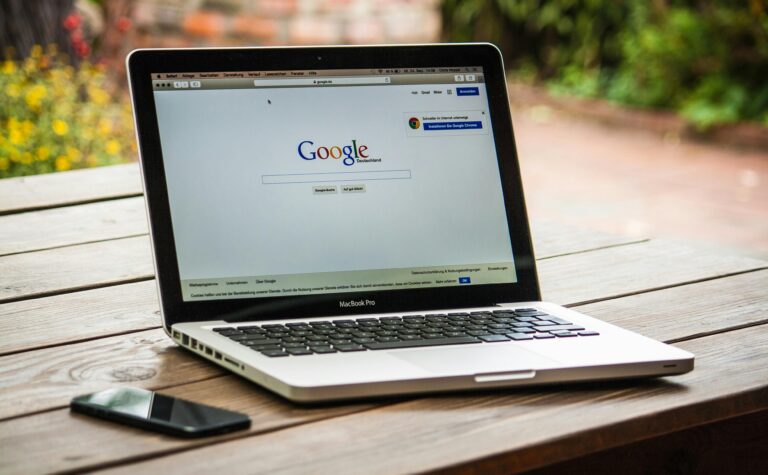 A MacBook Pro displaying Google Search on a wooden table outdoors, next to a smartphone.