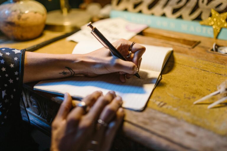 A hand with tattoos writing in a notebook on a rustic wooden desk, under warm lighting.